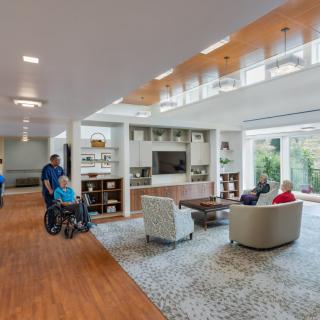 A spacious and well-lit community living area with seniors engaging in conversation and staff assisting in a care facility.