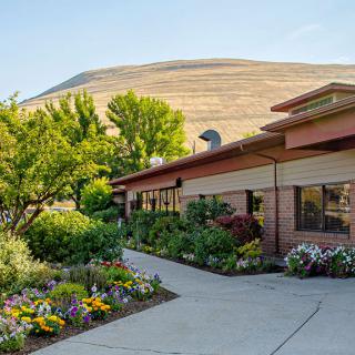 A building surrounded by a colorful garden with flowers and trees, set against a backdrop of hills.