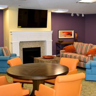 A vibrant living room with colorful furniture, featuring a striped sofa, orange and blue chairs, a fireplace topped with a TV, and artwork adorning the walls.