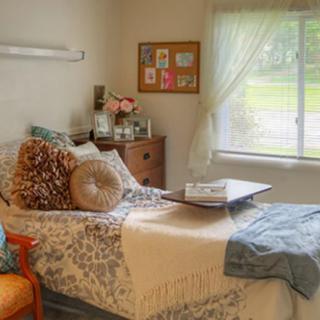 A cozy and well-decorated bedroom featuring a comfortable bed with patterned bedding, cushions, and a soft blanket beneath a sunlit window.