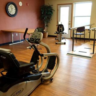A rehabilitation exercise room featuring fitness equipment including a stationary bike, with wood flooring and natural light.