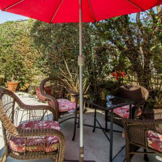 A cozy outdoor patio setup with wicker chairs, a table, and a red umbrella surrounded by lush greenery.