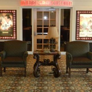 A cozy seating area in a movie theater lobby, featuring two armchairs and a table with a lamp, flanked by movie posters.