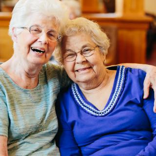 Two elderly women smiling and embracing indoors.