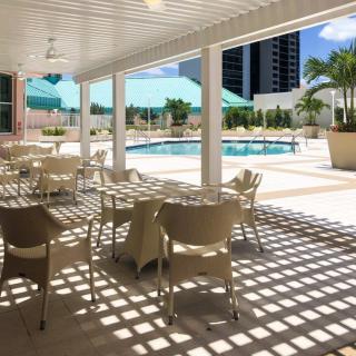 A sunny outdoor patio area with tables and chairs overlooks a swimming pool in a tropical setting.