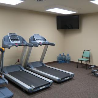 A small gym with two treadmills, a wall-mounted TV, several water bottles, and a chair.