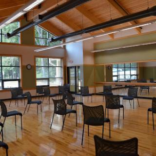 A spacious activity room with chairs arranged for gathering, featuring large windows that allow natural light to illuminate the wooden floor.