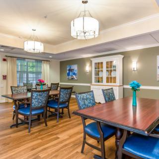 A well-lit dining area with wooden tables and blue chairs, featuring decorative elements such as flowers and artwork on the walls.