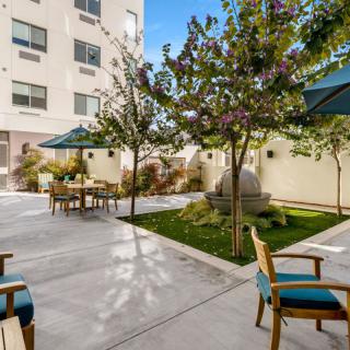 A serene courtyard with outdoor seating and umbrellas, surrounded by greenery and a multi-story building in the background.