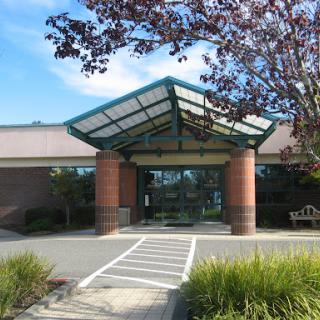 A modern healthcare facility entrance with a glass canopy and brick pillars, surrounded by landscaped greenery.