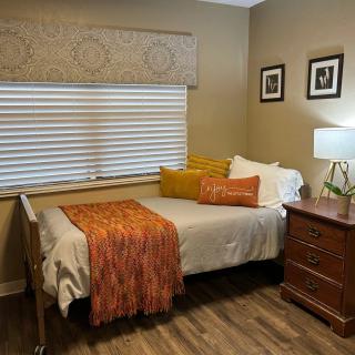 Cozy bedroom with a neatly made single bed, adorned with an orange throw blanket and decorative pillows. A nightstand with a lamp sits beside the bed, and framed pictures hang on the wall above.