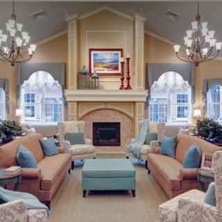 A cozy and elegantly designed living room featuring plush sofas, armchairs, a central fireplace, and large windows adorned with chandeliers.