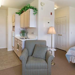 A cozy living space featuring a comfortable armchair, a kitchenette, and a neatly made bed.