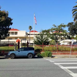 A view of a building with a flag and palm trees, featuring a blue vehicle parked nearby on a sunny day.