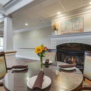 A cozy dining area featuring a table set for two with brown napkins, a bright flower vase centerpiece, and a warm fireplace in the background.