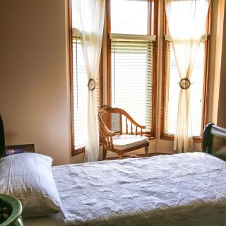 A cozy bedroom featuring natural light from large windows, a single bed, and a wooden chair.