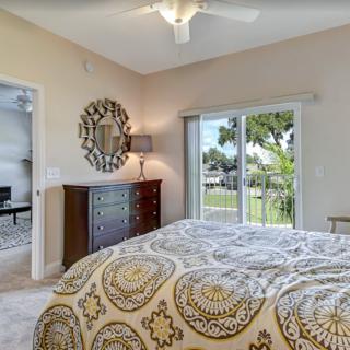 A cozy bedroom featuring a large bed with patterned covers, a decorative mirror above a wooden dresser, and large sliding doors opening to an outdoor view.
