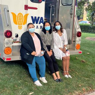 Three people wearing masks sit outside near a medical truck.