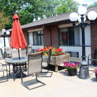 A cozy outdoor patio area with tables, chairs, and a red umbrella, surrounded by colorful flowers near a brick building.