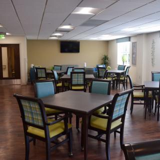 A spacious dining area with multiple tables and chairs, well-lit by ceiling lights and natural light from windows.