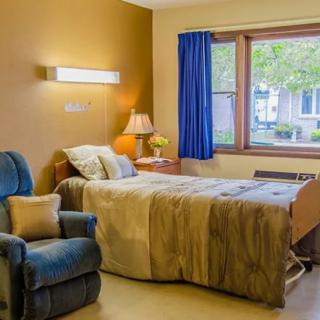 A cozy hospital room with a comfortable bed and chair, warm lighting, and a view through a large window.
