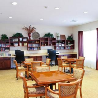 A well-lit community room featuring tables, chairs, computers, and bookshelves adorned with decorative plants.