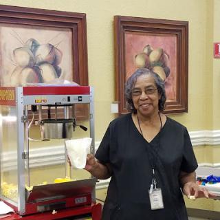 A woman stands beside a popcorn machine, ready to serve snacks at an indoor event.