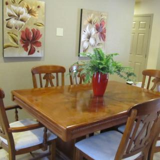 A cozy dining room with a wooden table, surrounded by chairs, adorned with a red vase and greenery. Floral paintings hang on the wall.