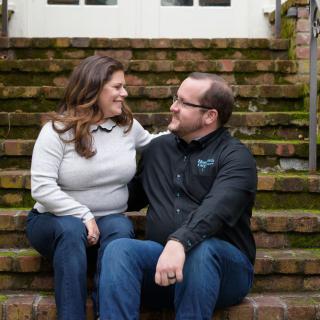 A couple sitting on outdoor stairs, smiling at each other in casual attire.
