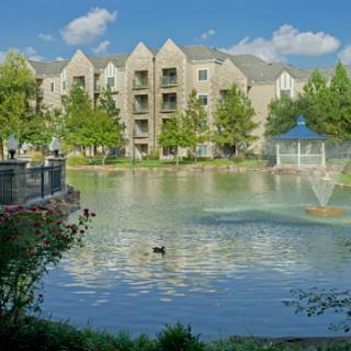A serene view of an apartment complex by a tranquil lake, featuring lush greenery and a fountain.