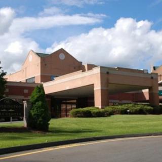 A brick building that appears to be a hospital, surrounded by trees and landscaping under a cloudy sky.