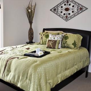 A cozy bedroom with a neatly made bed featuring green bedding and decorative pillows, complemented by wall art and a vase in the corner.