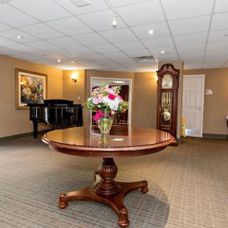 A spacious room featuring a round wooden table with a flower arrangement, a grandfather clock, and a piano in the corner.
