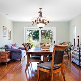 A well-lit dining room with a wooden table and chairs, featuring a chandelier and natural light from large windows.