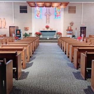 An interior view of a church featuring wooden pews leading up to an altar with a cross and colorful stained glass above.