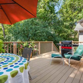 A sunny deck with a table, umbrella, lounge chairs, and a grill surrounded by trees.