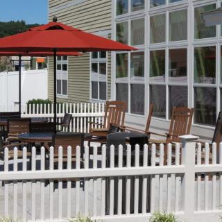 An outdoor patio area with wooden chairs and a table under a red umbrella, surrounded by a white picket fence.
