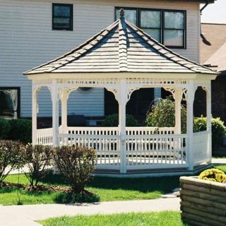 A wooden gazebo situated in a sunny yard with manicured lawn and shrubs.