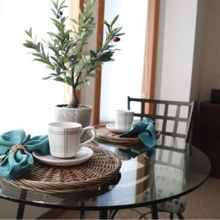A cozy dining setup with a glass table featuring white tea cups, blue napkins, and a potted plant centerpiece.