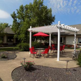 A cozy outdoor patio with red chairs under a white pergola and umbrella on a sunny day.