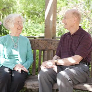 An elderly couple sitting on a bench outdoors, enjoying a conversation on a sunny day.