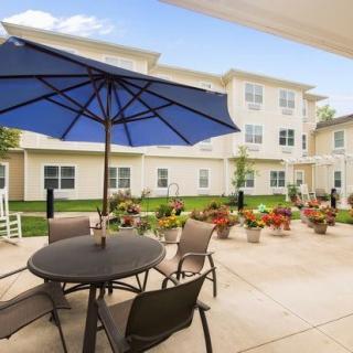 A sunny outdoor patio with a table, chairs, and a large blue umbrella, set against a backdrop of a multi-story residential building with a garden.