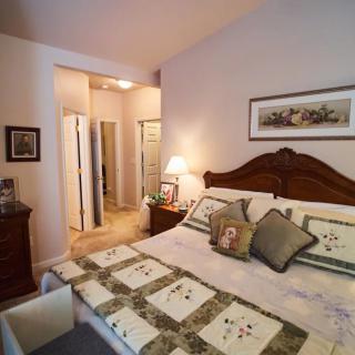 A cozy bedroom with a wooden bed frame, decorative quilt, and matching pillows. A bedside lamp illuminates the room, complemented by artwork on the wall.
