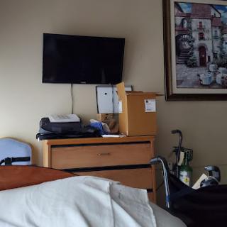 A view of a hospital room featuring a TV, wooden furniture, and a piece of artwork on the wall.