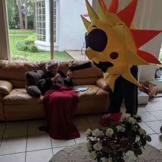 A person in a sun costume interacts with someone sitting on a sofa in a living room.