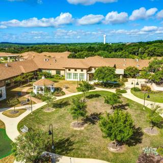 Aerial view of a large building complex surrounded by lush greenery and winding pathways under a bright blue sky.