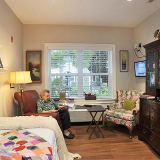 An elderly person sits comfortably in a cozy living room adorned with colorful quilts and a window view.
