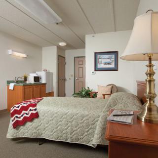 A cozy hotel room featuring a neatly made bed, bedside table with a lamp, and a small kitchenette in the corner.