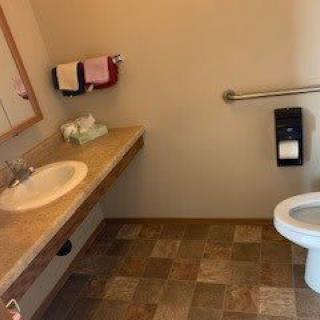 A bathroom with a sink, mirror, and toilet, featuring tiled flooring and a handrail.