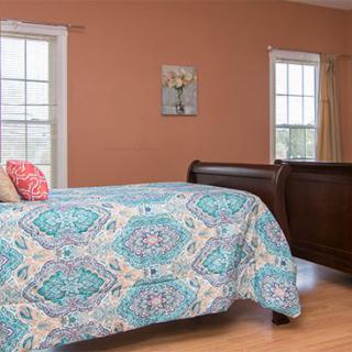 A cozy bedroom with a patterned quilt and warm-toned walls, featuring two windows and decorative wall art.